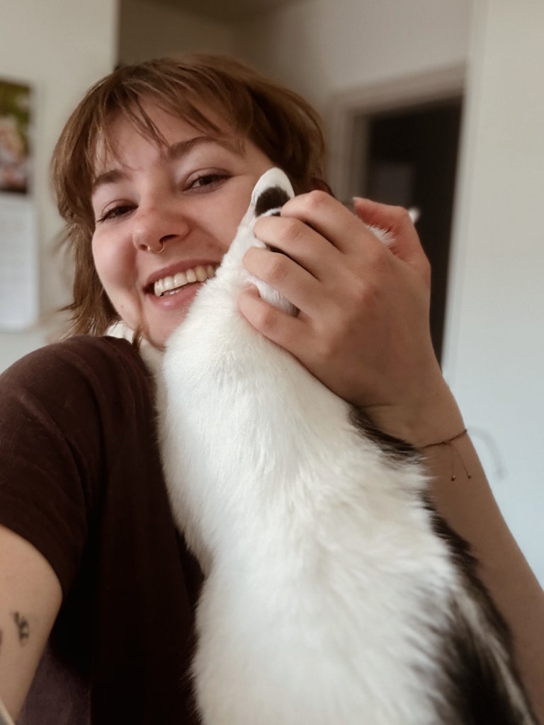 Uśmiechnięta kobieta przytulająca się z białym kotem w czarne łatki. Kompleksowa i czuła opieka dla kotów z różnymi potrzebami.