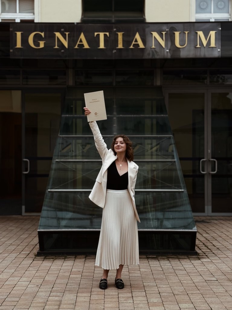 Uśmiechnięta kobieta Izabela Stańczyk trzymająca pracę magisterską z psychologii na tle budynku Uniwersytetu Ignatianum po udanej obronie. Solidne wykształcenie psychologiczne jako fundament profesjonalnego wsparcia psychologicznego w Krakowie dla dzieci i dorosłych.