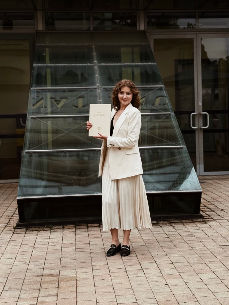 Uśmiechnięta kobieta w kremowej marynarce i spódnicy plisowanej stoi przed Uniwersytetem Ignatianum w Krakowie, trzymając w rękach pracę z obrony magisterskiej. Psycholog Kraków dla dzieci, dorosłych i seniorów.