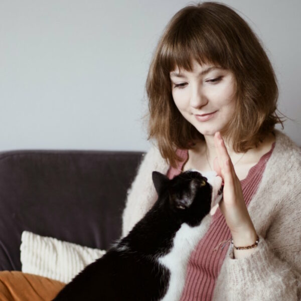 Izabela Stańczyk siedzi na szarej kanapie i trzyma prosto rękę z przysmakiem rozsmarowanym na wnętrzy dłoni, który zlizuje czarno-biały kot. Dietetyk i behawiorysta kotów w Krakowie i online.