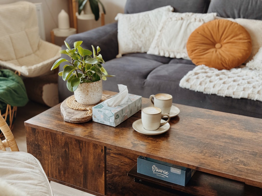 Drewniany stolik kawowy z dwiema filiżankami kawy, pudełkiem chusteczek i rośliną doniczkową na tle szarej kanapy. Wsparcie psychologiczne dla dzieci i dorosłych w trudnych chwilach i żałobie.